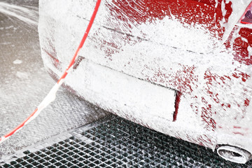 Red car washed in self serve carwash, detail on soap foam spraying from high pressure hose back,...