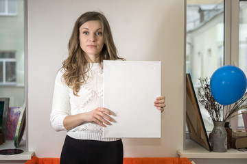 A woman stands indoors between windows and holds an empty sheet of paper with copy space for your...