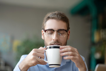 Young man with glasses looking smiling at a cup of coffee.