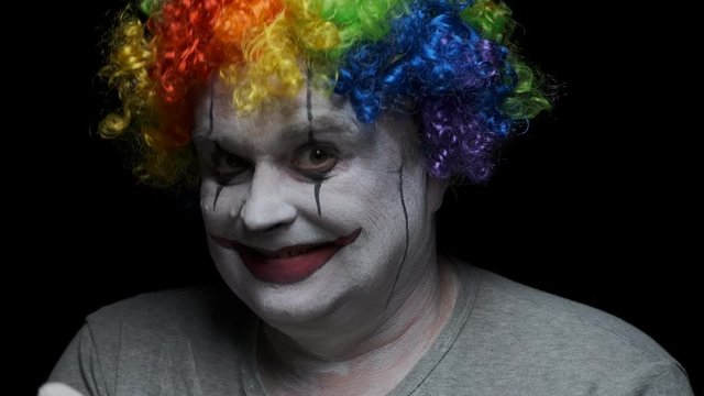 Portrait of smiling clown man inviting victim with hand on black background.