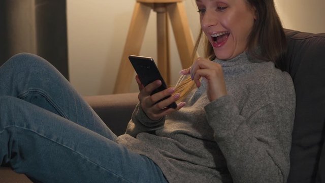 Smiling woman lying on the couch in a cozy room and using smartphone for surfing internet or chatting with someone in the evening. She is very happy that she sees