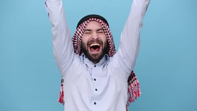 Bearded young arabian muslim man 20s in keffiyeh kafiya ring igal agal casual clothes doing winner gesture, say Yes raise hands up isolated on pastel blue background People religious lifestyle concept