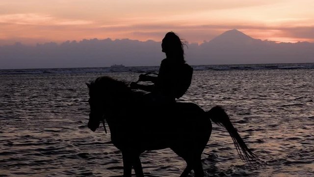 Silhouette slow motion shot of man riding a horse during sunset at the beach of Gili Trawangan, Indonesia. Ocean and active volcano Mount (Gunung) Agung in Bali covered in clouds in the background.