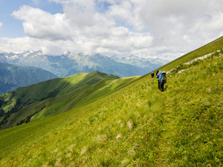 Fototapeta na wymiar Hiker with a backpack goes on the grassy slope on a background of mountains. Back view. Beautiful sunny day in Arkhyz. Concept of healthy lifestyle, trekking activity, hiking adventure