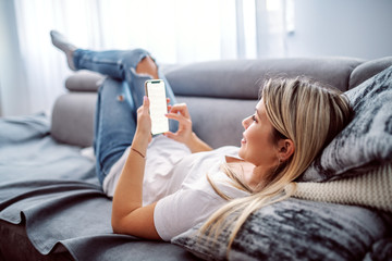 Charming caucasian blond young woman dressed casual lying down on sofa in living room, relaxing and using smart phone.
