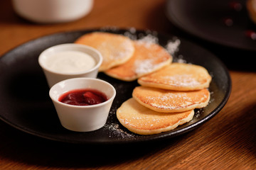 Cooking pancakes for breakfast. Small round fritter with sour cream and berry jam and black tea cup on the background, close up.