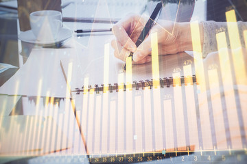 Hand taking notes in notepad. Forex chart holograms in front. Concept of research. Double exposure