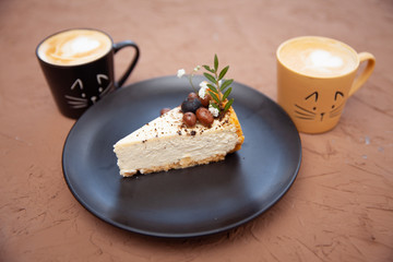 Two cups with a coffee stand near a black plate on which lies a piece of cake on which there are nuts and fruits
