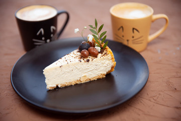 Two cups with a coffee stand near a black plate on which lies a piece of cake on which there are nuts and fruits