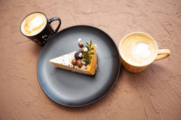 Two cups with a coffee stand near a black plate on which lies a piece of cake on which there are nuts and fruits