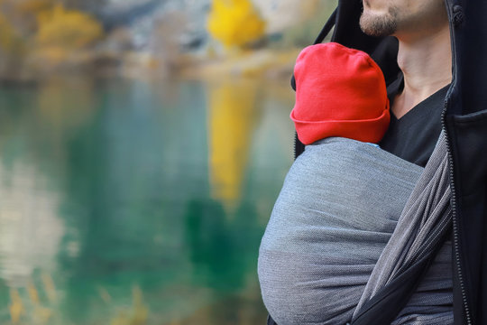 Attractive young father with her infant baby in sling outdoor. Man is carrying her child and travel in autumn mountain lake. Babywearing concept