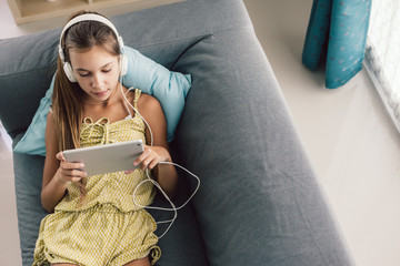 Pre teen child playing tablet and listening to music in head phones while relaxing on couch in living room at home