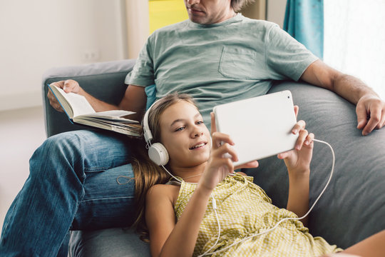 Father and pre teen daughter playing tablet and reading book while relaxing together on couch in room at home