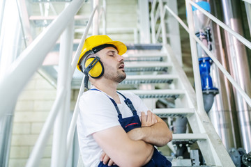 Overworked caucasian worker in overall, with hardhat and antiphons leaning on railing with legs...