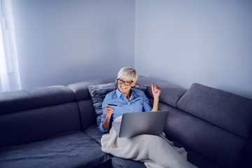 Cheerful charming caucasian senior blond woman with short hair and with eyeglasses sitting on sofa in living room, holding laptop in lap and using credit card for online shopping.