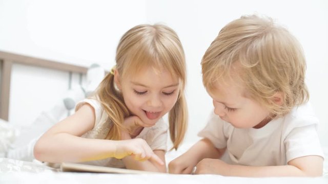 Cute blond kids play games on tablet in a light room.