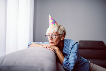 Sad caucasian blond senior woman with birthday cap on head sitting on sofa in living room and leaning.