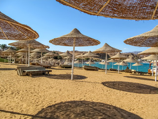 Hotel beach without people in the bay of the Red Sea in Sharm El Sheikh (Egypt). Solar umbrellas with thatched roofs on yellow sand on a background of blue water on a sunny summer day