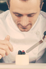 Smiling chef decorating ice-cream with berries. Man holding spatula and putting blueberries and raspberry on piece of ice-cream at table. Dessert and culinary concept. Front view.