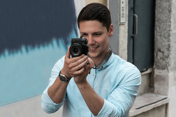 Smiling young man taking photo on camera in street. Handsome guy standing with building wall and door in background. Tourism and leisure concept. Front view.