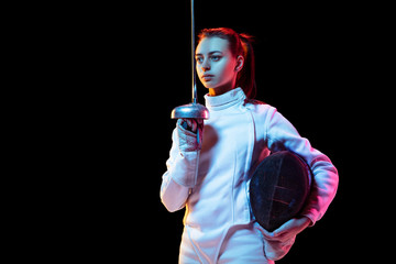 Confident. Teen girl in fencing costume with sword in hand isolated on black background, neon light. Young model practicing and training in motion, action. Copyspace. Sport, youth, healthy lifestyle.