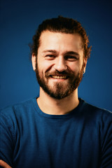 Obraz premium Young charming attractive Caucasian bearded hipster with curly hair dressed in sweater standing in front of blue background and looking at camera.