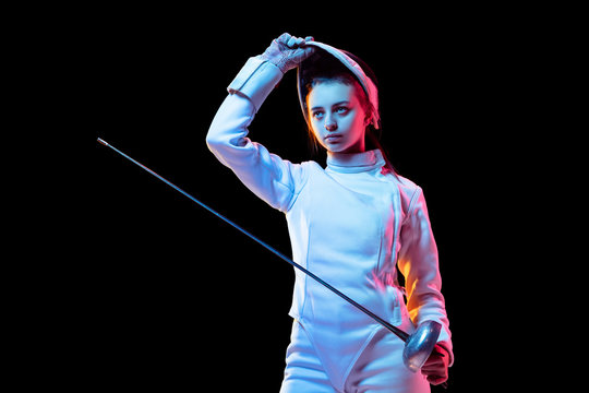 Ready. Teen girl in fencing costume with sword in hand isolated on black background, neon light. Young model practicing and training in motion, action. Copyspace. Sport, youth, healthy lifestyle.