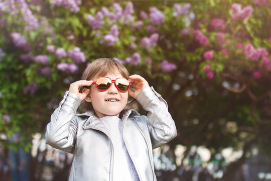 little girl in red sunglasses in silver jacket smiles and walks in flowering park in spring on the background of flowering trees