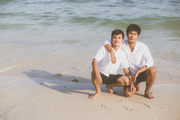 Fototapeta premium Young asian couple gay smiling romantic drawing heart shape together on sand in vacation, homosexual happy and fun hug with love sitting on sand at the beach in travel summer, LGBT legal concept.