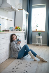 Dark-haired girl in a grey sweater sitting on the floor and having coffee