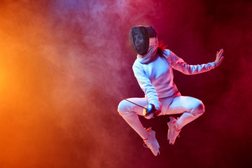 Fototapeta na wymiar Blooming. Teen girl in fencing costume with sword in hand isolated on black background, neon lighted smoke. Practicing and training in motion, action. Copyspace. Sport, youth, healthy lifestyle.