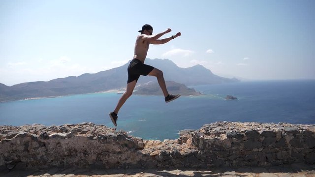 Slowmotion of Young Man Jumping on Old Stone Wall With Greek Island in Background. Summer Vacation Fun Concept