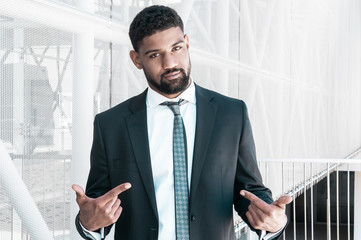 Confident black business man pointing at himself. Serious guy looking at camera and standing with building constructions in background. Company representative concept. Front view.
