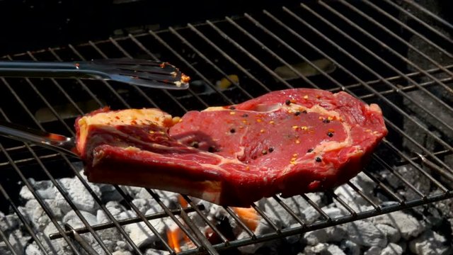 Piece of meat steak sprinkled with pepper and spices placed on the grill with meat tongs