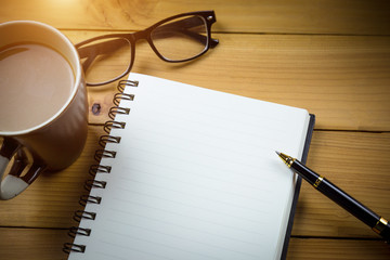 Blank notebook with pen and with glasses next to cup of coffee on wooden table,business concept.