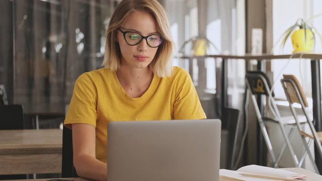 A concentrated blonde woman wearing eyeglasses and yellow t-shirt is working on her laptop while sitting in a conference hall