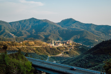 Summer landscape from the inside of Liguria, Italy. Bridge over a green valley, mountain hills with dense forests, typical small Italian village with colorful houses, narrow winding roads.