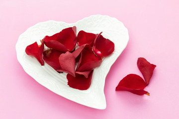 Separated petals of red roses on white plate in the shape of heart. Red rose petals on white plate on pink background.