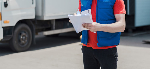 Courier driver in uniform making notes in document and delivery white truck behinde him