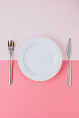 Empty white plate with cutlery on a pink-peach background. Top view. Vertical
