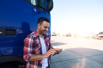 Professional smiling truck driver checking using tablet computer and standing by long vehicle....
