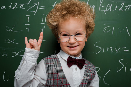 happy and smart child in suit and bow tie showing rock sign near chalkboard with mathematical formulas