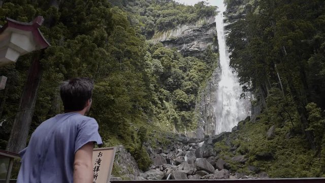 Man walking up to Nachi Falls in Nachikatsuura, Japan; huge flowing waterfall in background, slow motion