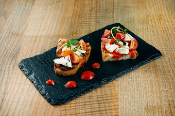 Traditional Italian antipasti - bruschetta with feta and tomatoes on a black slate board on a wooden background.