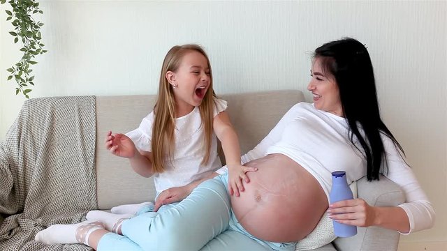 Young pregnant mother and her daughter moisturizing mom`s stomach with cream. Little girl applying body lotion on her mother`s stomach.