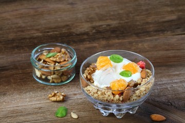 Breakfast cereals in a bowl.  Fresh granola, muesli  with yogurt, tangerine, almonds, walnuts and ice cube on wooden table. Basil leaves.