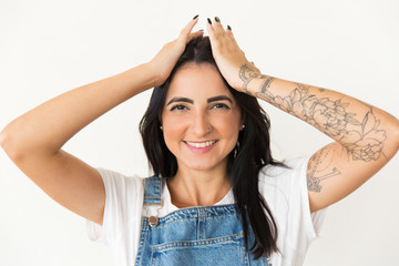 Fototapeta premium Cheerful young woman smiling at camera. Beautiful happy young tattooed woman holding hands on head and looking at camera isolated on white background. Happiness concept