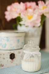White natural candle in vintage glass jar with lace ribbon. Gift boxes with flower ornament. Bouquet of tulips with pink and white petals in white metal vase.