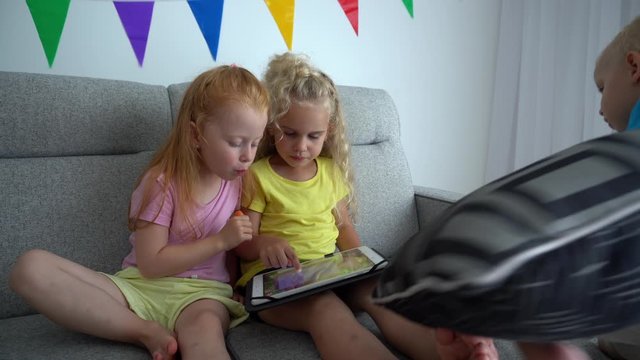 Redhead girl eat carrot and blond one using tablet. Little brother sit on sofa