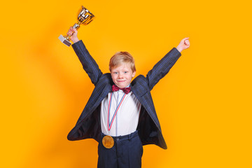 Cheerful boy holding a golden trophy and gold medal on yellow background - Powered by Adobe